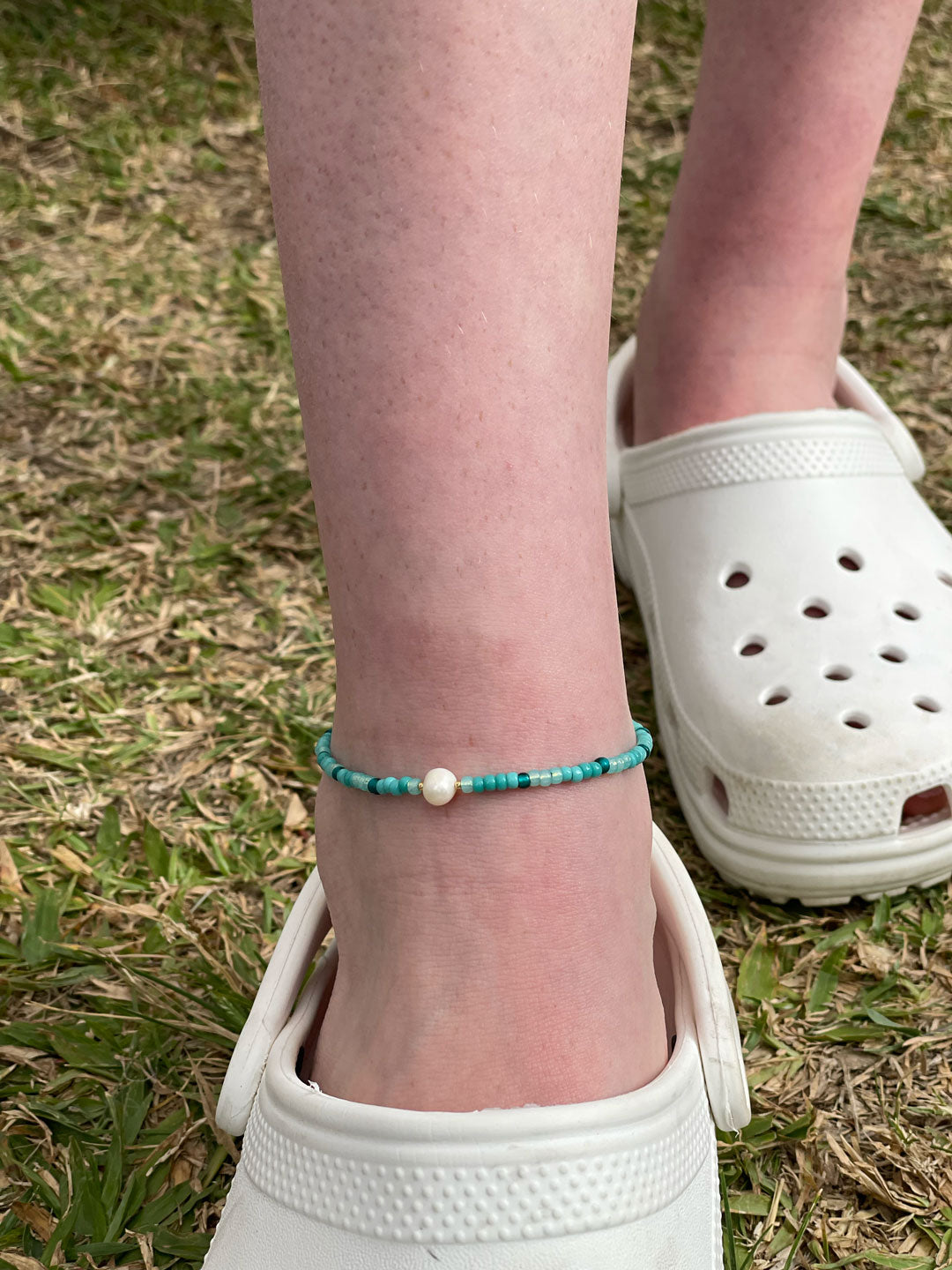 Person wearing a turquoise beaded anklet with a white croc shoe on grass.