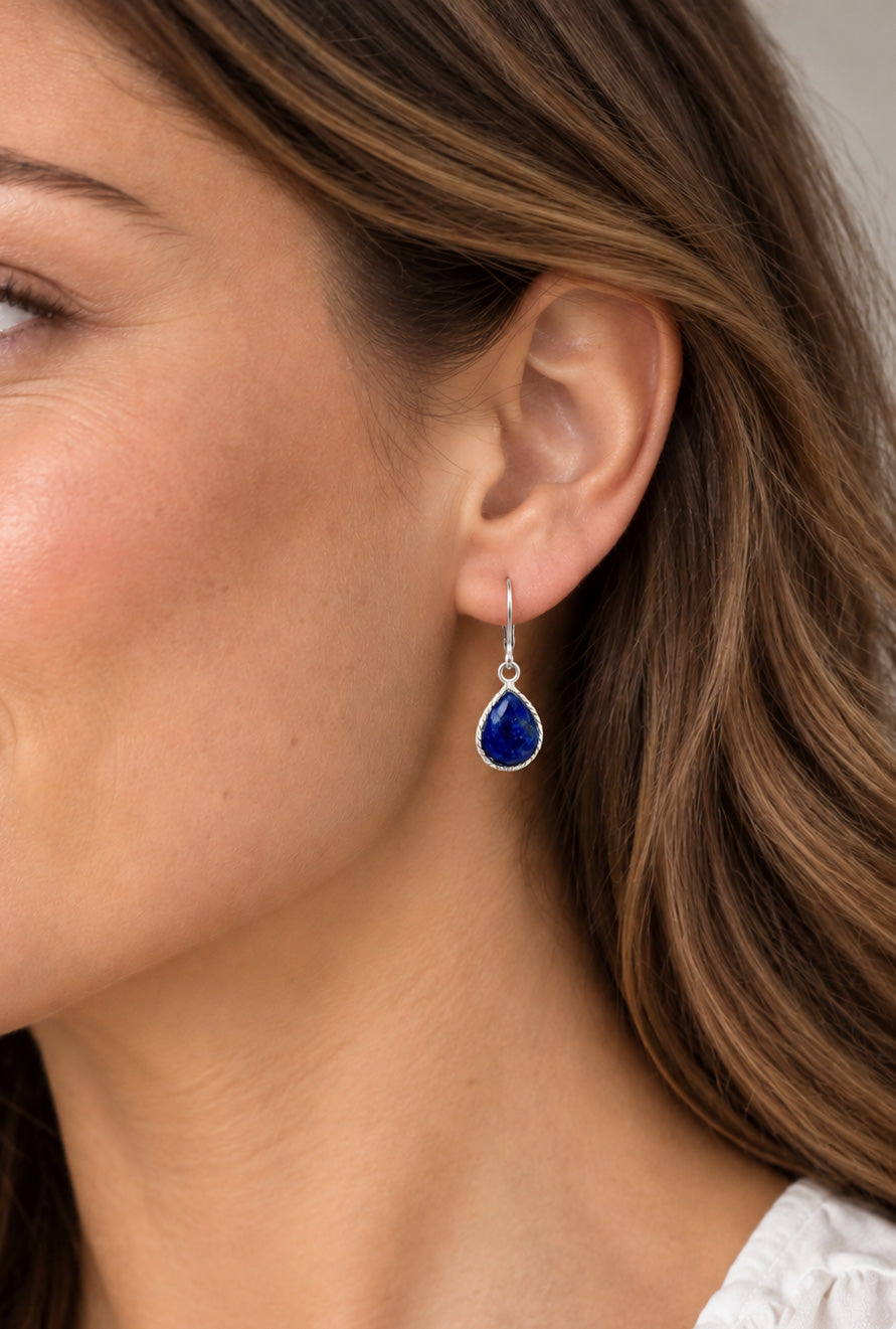 A close-up of a woman's ear wearing a single teardrop-shaped blue gemstone dangle earring with a silver-toned setting and hook.