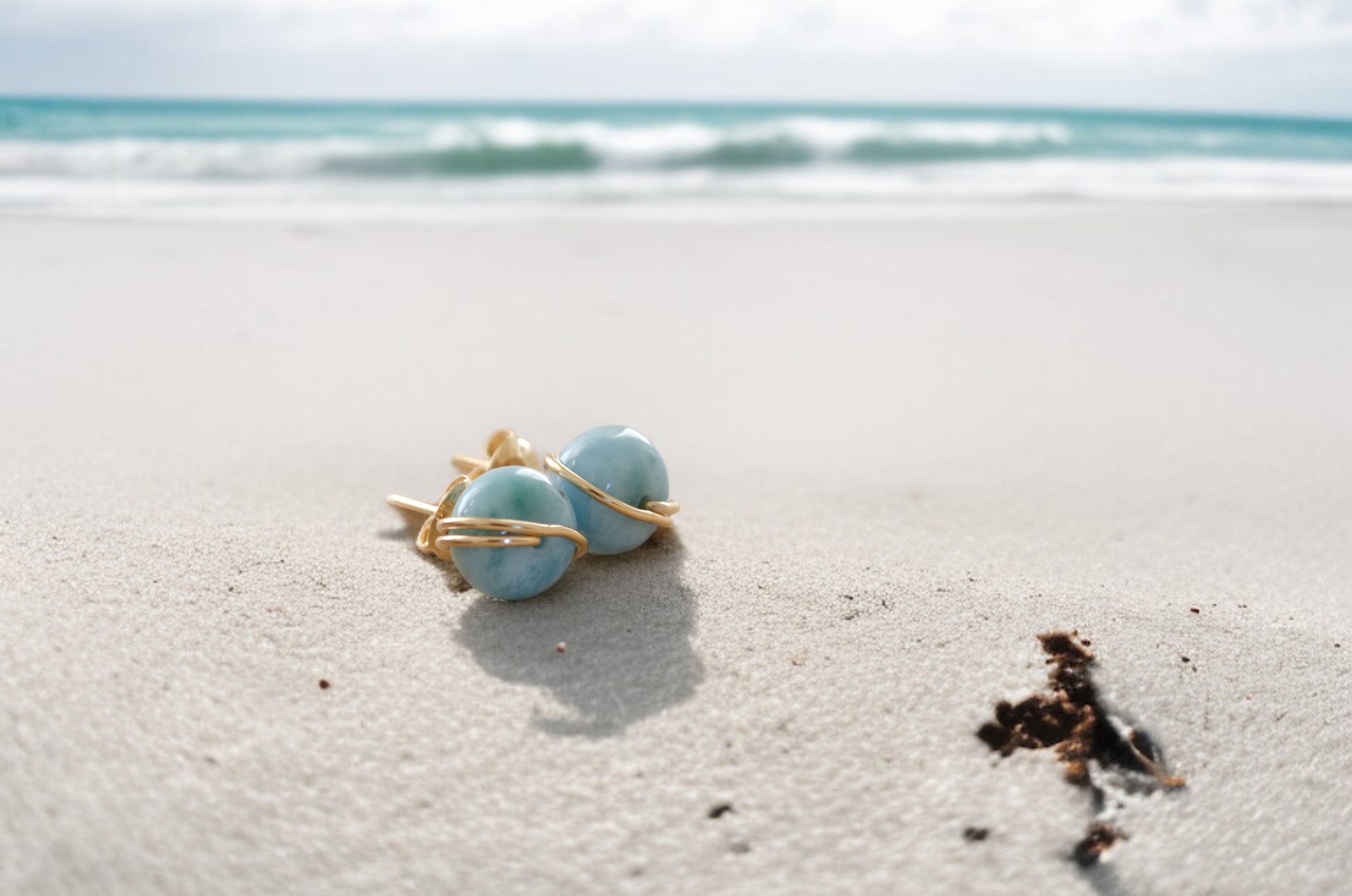 Pair of Larimar earrings on a sandy beach with ocean in the background