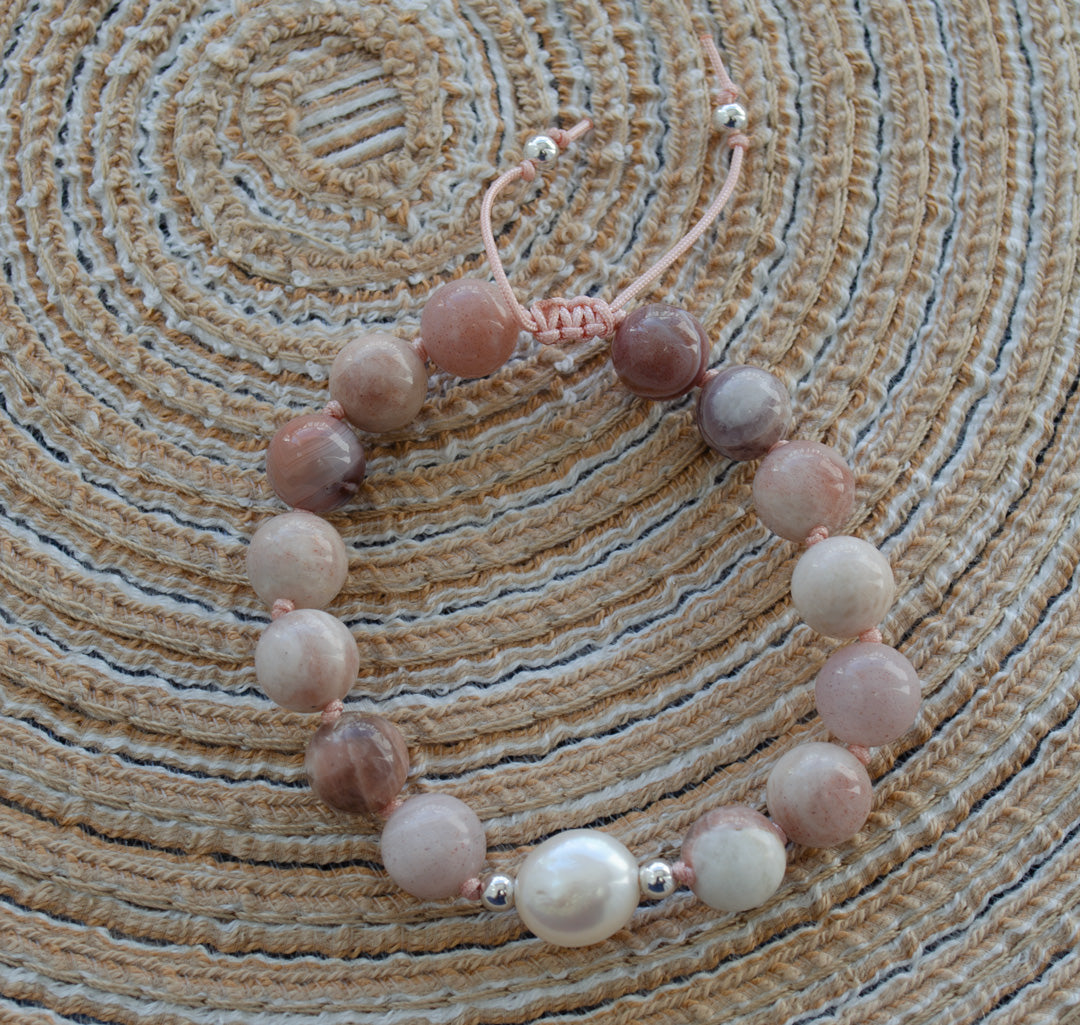 A beaded bracelet with round pink, mauve, and white beads, featuring a central white pearl and small silver spacer beads, resting on a woven straw mat.