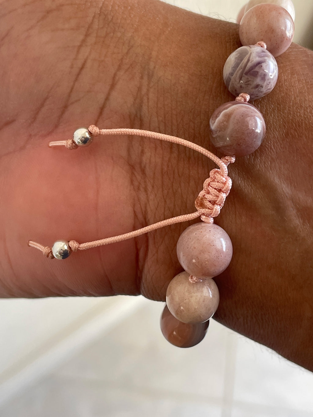 A close-up of a beaded bracelet worn on a wrist. The bracelet features smooth, polished round beads in dusty pink, mauve, and light brown tones, with a peach cord and small metallic silver beads.