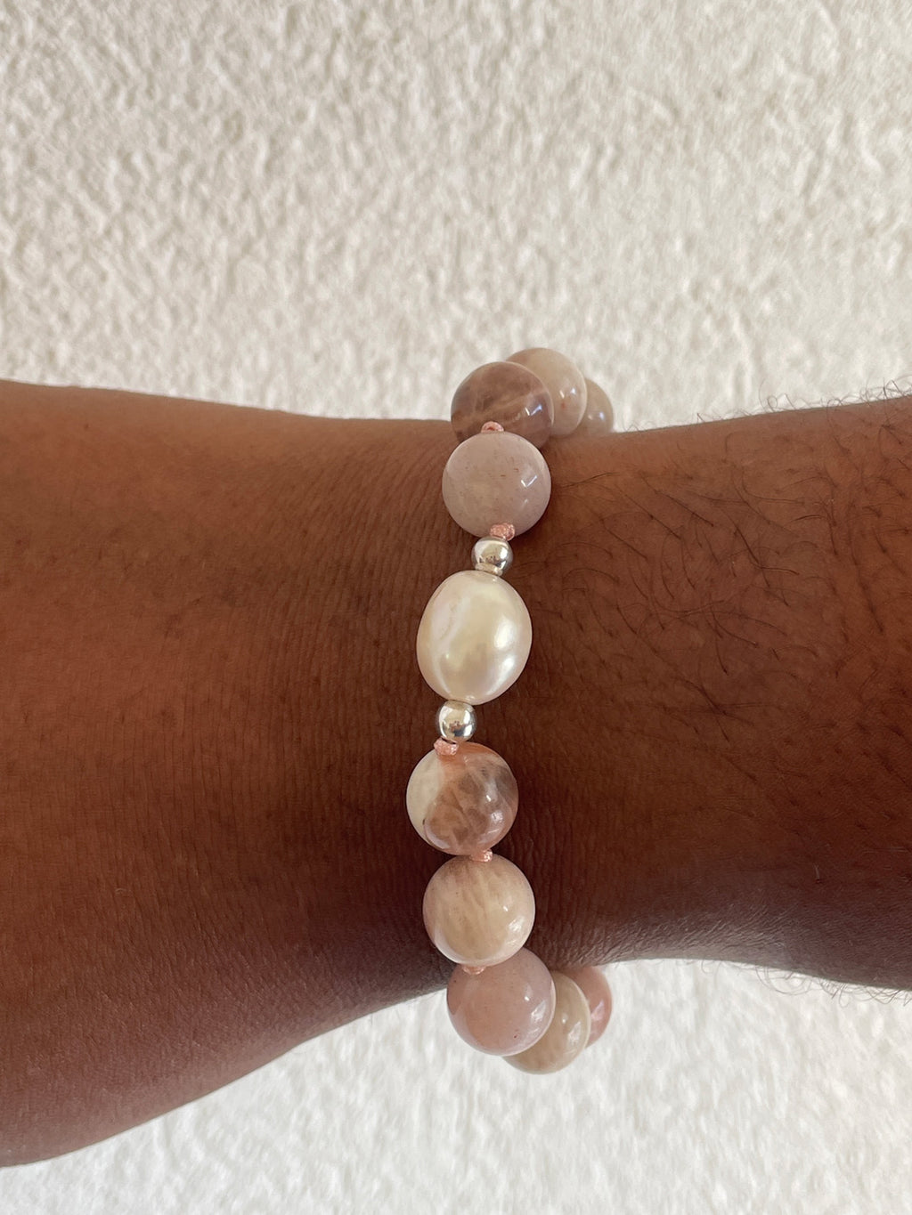 A beaded stretch bracelet with pink rose quartz and white baroque pearl beads, shown worn on a person's wrist against a textured off-white wall.