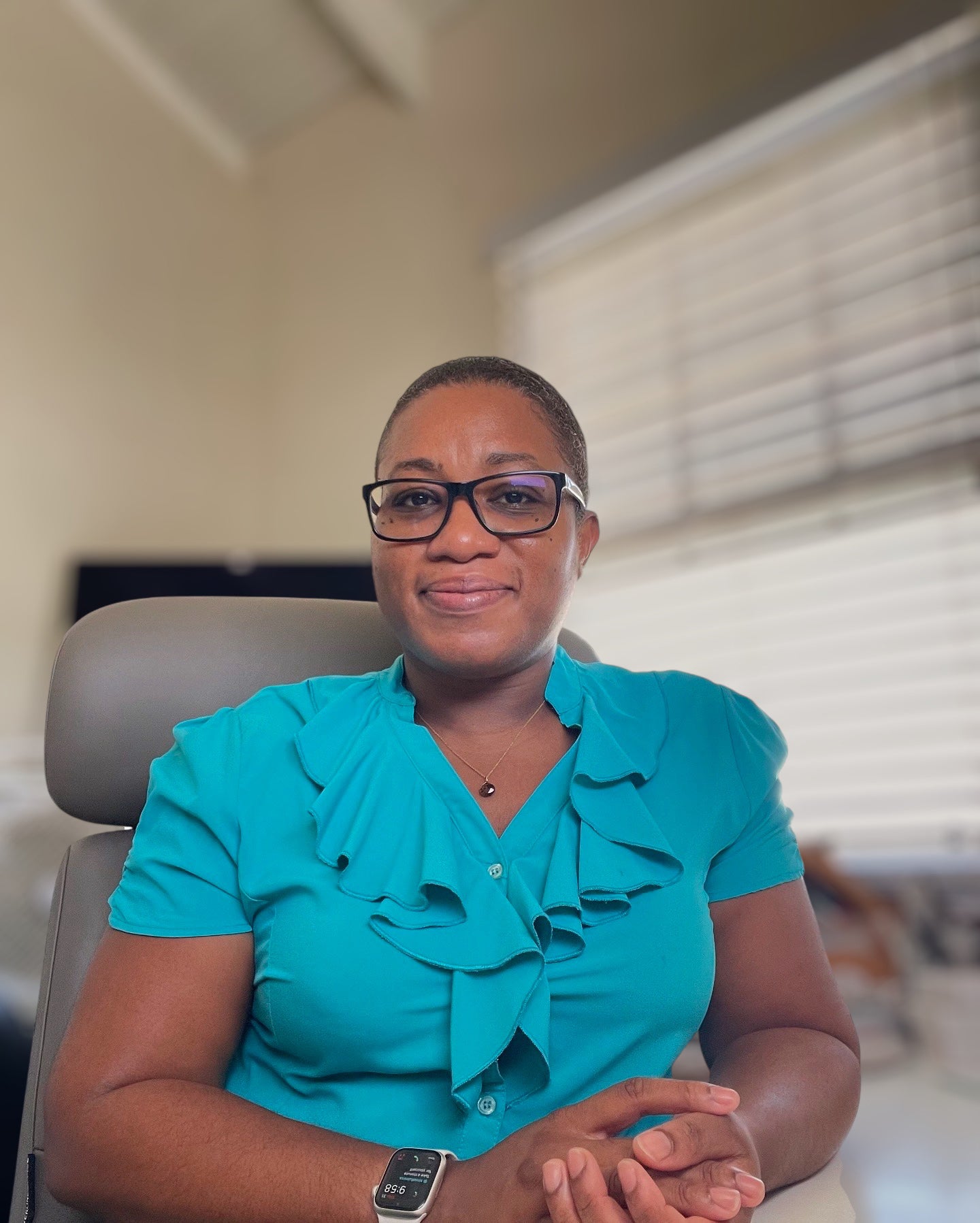 Woman wearing glasses and a teal blouse sitting in an office chair.