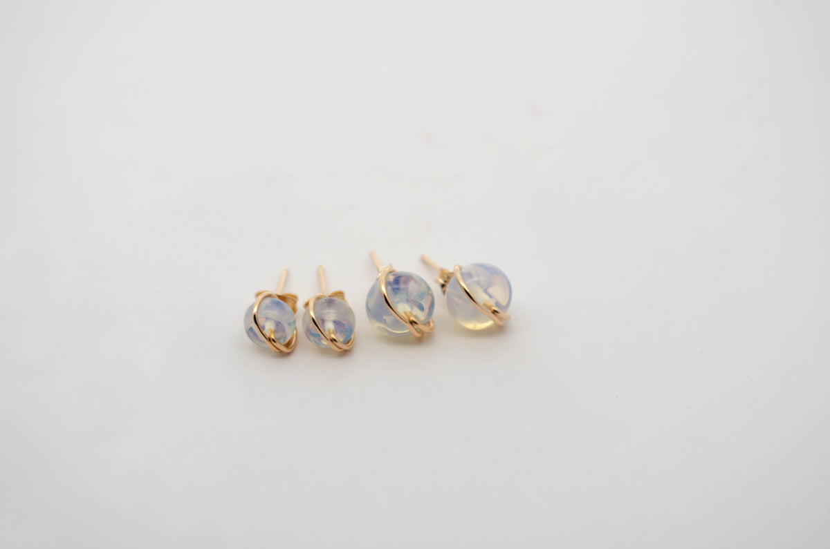 Three pairs of gold stud earrings with round, milky white gemstones, arranged on a plain white background.